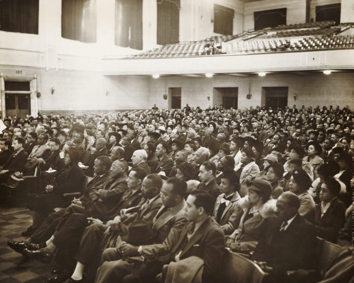 A large audience of mostly Black men and women in formal attire sit in an auditorium, attending a Brotherhood of Sleeping Car Porters event.