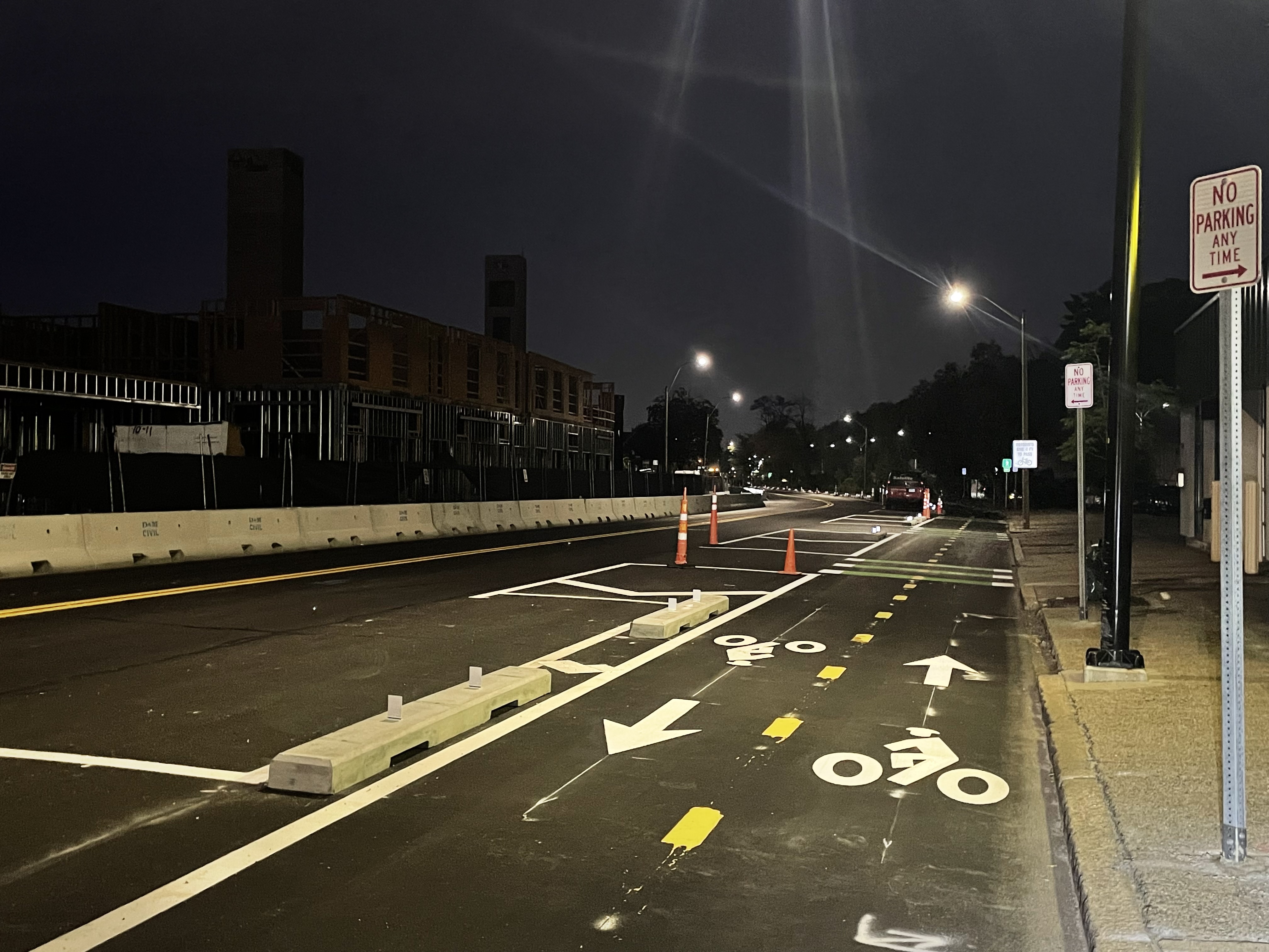 A city street pictured in the evening after sundown. In the foreground is a two-way bike path, separated from the rest of the street by concrete curb barriers. On the opposite side of the street is a construction site.