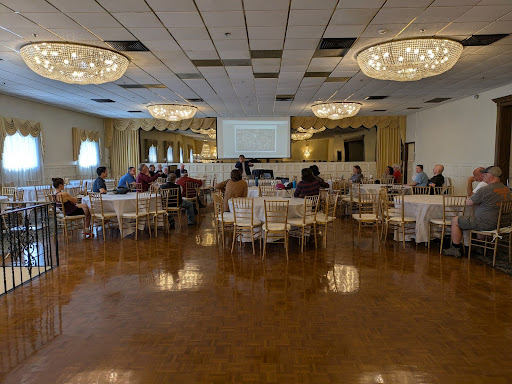 A community meeting in a banquet hall with chandeliers. About two dozen people sit at round tables listening to a speaker in front of a screen.