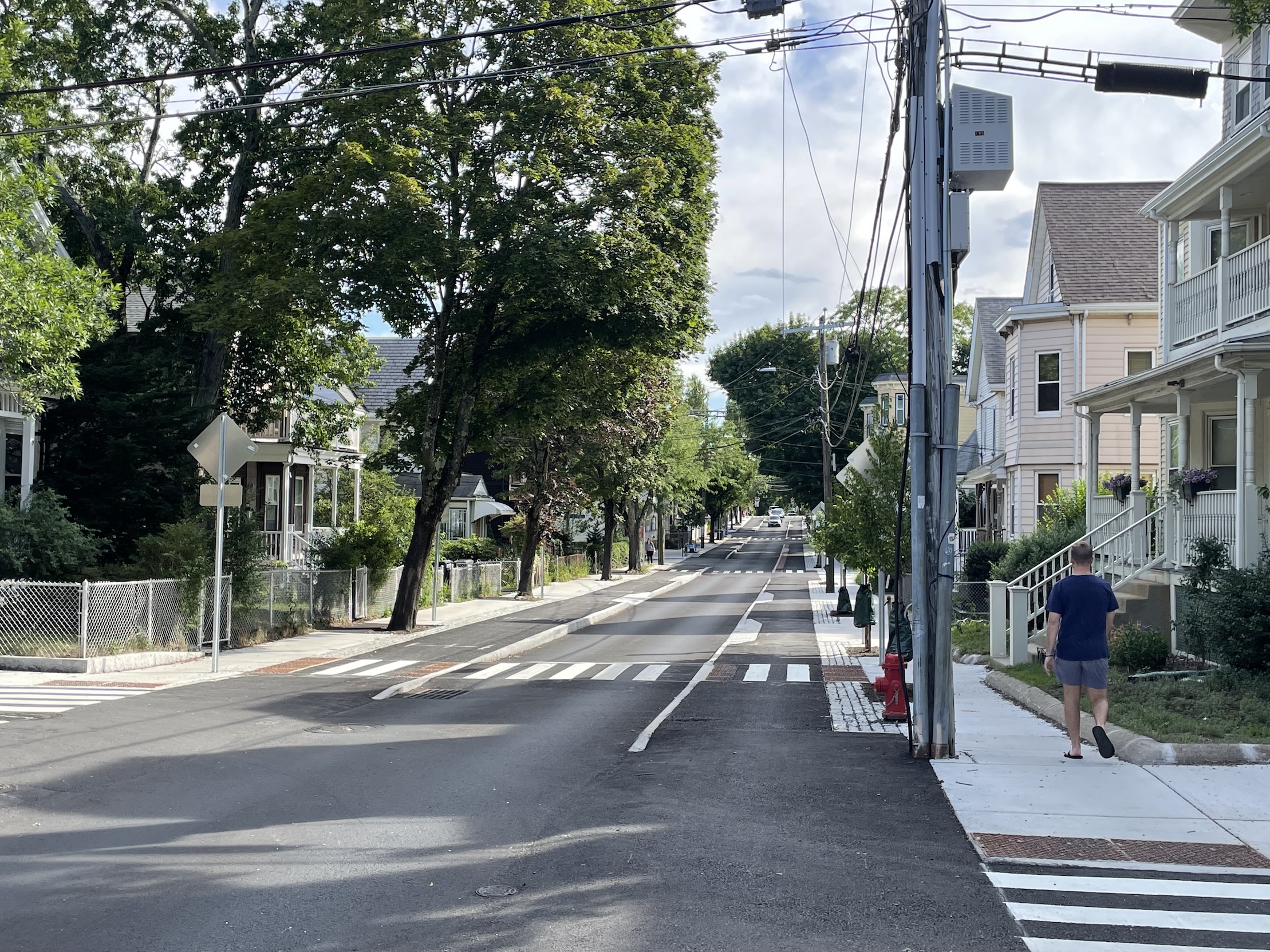A city street lined with small 3-story multi-family residential buildings. Two concrete curbs in the middle of the street divide a single lane of motor vehicle traffic from two sidewalk-level bike lanes on either side. White zebra-striped crosswalks cross the street every block into the distance. The street is also lined with leafy trees of varying sizes and ages, some of which are very large and mature, others of which have just been planted.