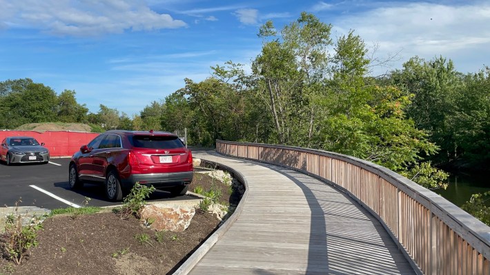A wooden boardwalk with a wooden railing to the right curves around a parking lot with two parked cars at left. At the far right edge of the photo is a river lined with trees.