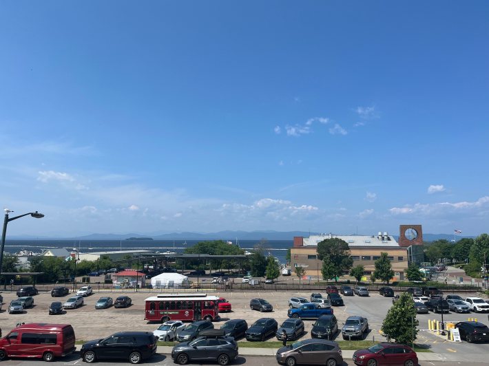 A view over a parking lot and trolley shows Lake Champlain, the mountains beyond it, and a clear, blue sky.