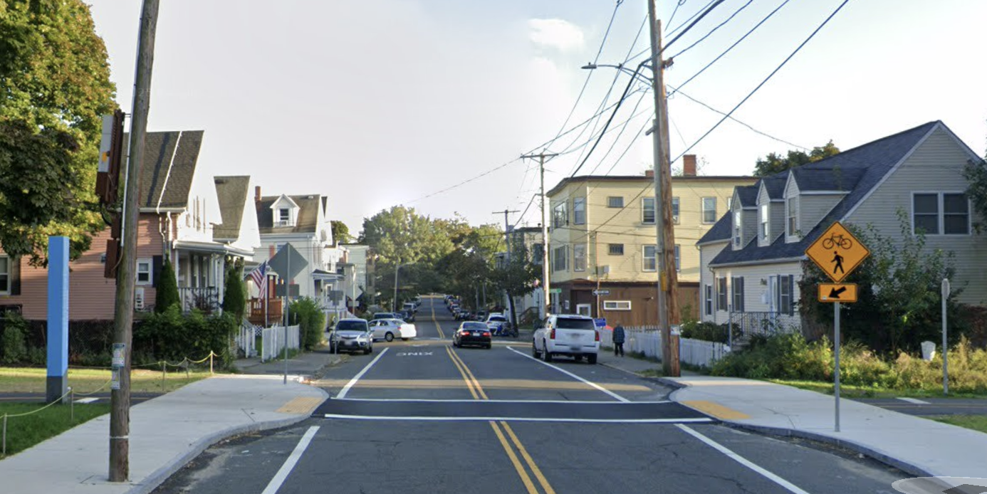 A crosswalk for a bike path crosses a two-lane city street lined with 2- and 3-story residential buildings next to a yellow diamond pedestrian crossing warning sign.