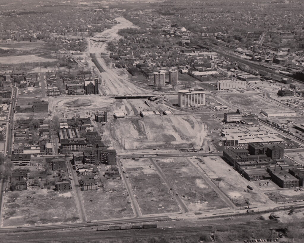 A black-and-white bird's eye view photo of a city with a swath of empty, bulldozed land in the foreground and in a long corridor leading towards the horizon. 