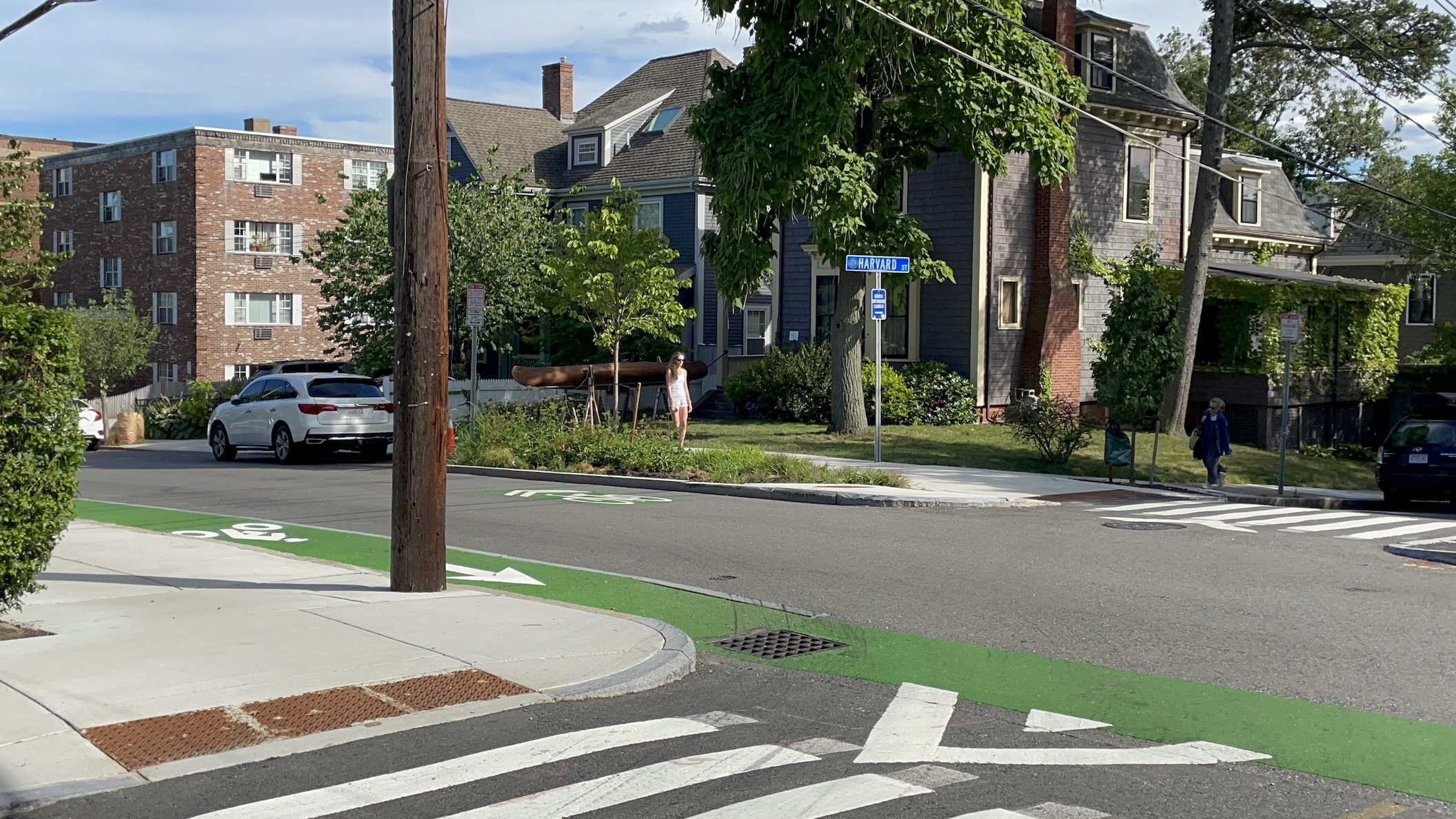 A residential city street lined with multi-story apartment buildings. A blue street sign in the center of the photo at an intersection on the opposite side of the street says "HARVARD ST." In the foreground a green-painted bike lane runs along the near side of the street from the left edge of the photo to the lower right corner. A garden with trees and shrubs lies between the street and the sidewalk on the opposite side of the street.