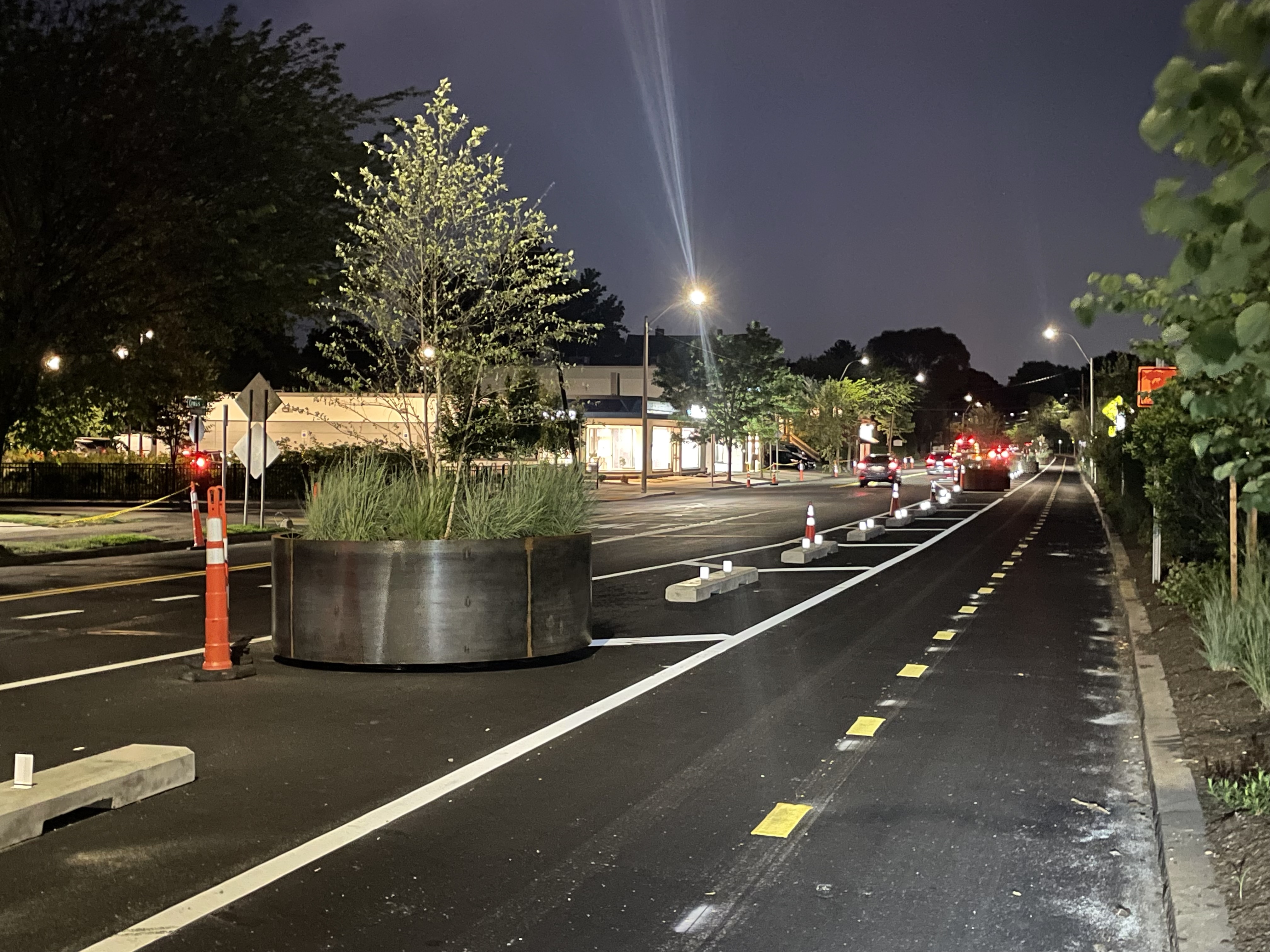 A city street pictured in the evening after sundown. In the foreground is a two-way bike path, separated from the rest of the street by concrete curb barriers and a large circular planter with a tree.