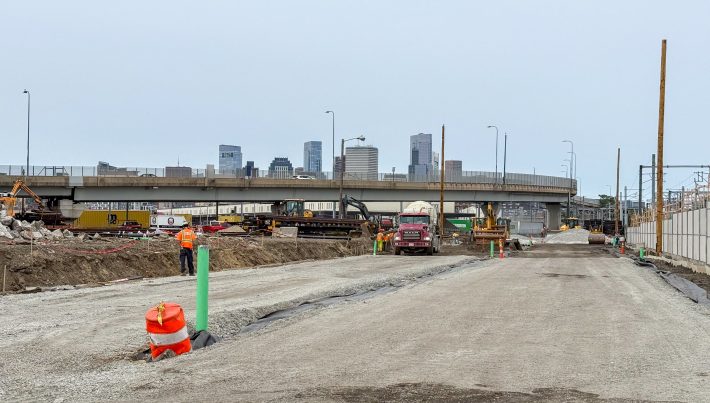 Construction workers work alongside heavy equipment on a broad, flat field of gravel with a concrete overpass in the distance and the Boston sky line beyond.