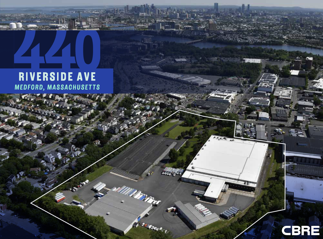 An aerial view of a cluster of three warehouses surrounded by smaller homes in a dense urban neighborhood. In the distance is the Boston city skyline on the horizon. A white line over the photo outlines the boundaries of the land the warehouses sit on and a blue and white title gives the property address as 440 Riverside Ave., Medford, MA.