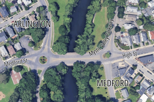 A satellite view of two traffic circles on either side of a river that runs vertically through the center of the image. The left side is labelled "ARLINGTON" and the right is "MEDFORD". A street labeled Mystic Valley Parkway connects the two traffic circles and continues vertically along the river; to the right, a street labelled High St. enters the right rotary from the right edge, and to the left, a street labelled Medford St. enters the left rotary from the Arlington side. 