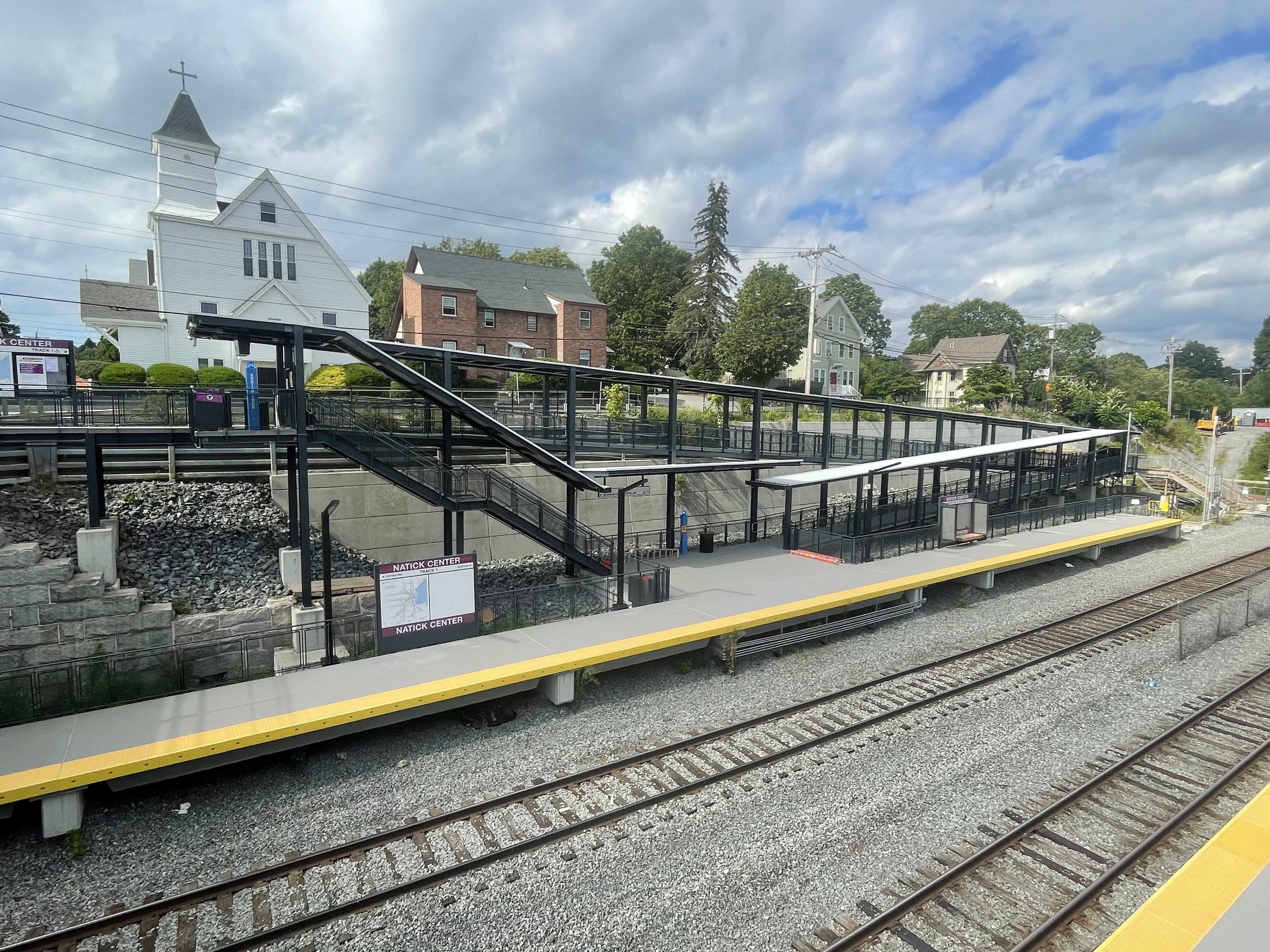 An elevated view of a commuter rail station across two railroad tracks. A sign on the platform reads "NATICK CENTER"
