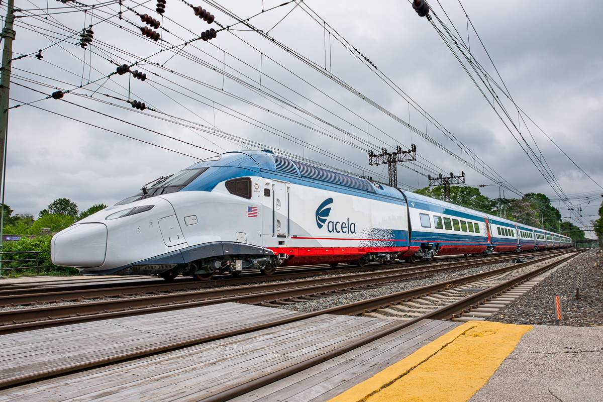 A blue-and-white Acela high-speed train, shot from a low angle near an empty set of railroad tracks in the foreground and overhead wires above.