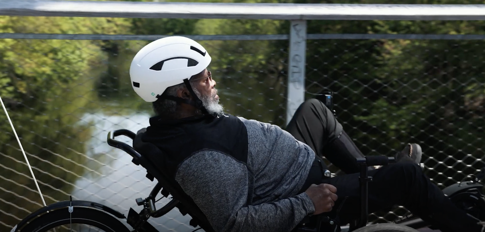 An older Black man with a white beard wearing a sweatshirt and a white bike helmet pedals a recumbent bike on a bridge overlooking a river.
