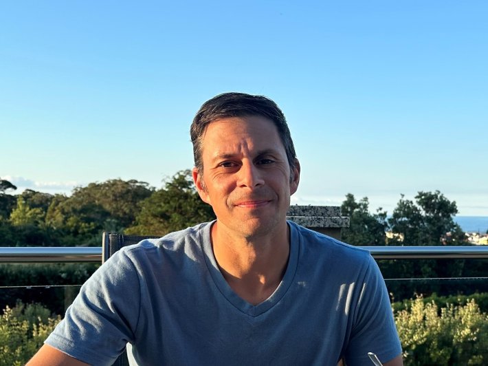 A photo of a man grinning in front of a field in the summer time.