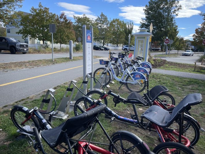A row of 7 bikes parked on a grassy area next to a paved bike trail. The three bikes in the foreground are adaptive recumbent tricycles and four bikes in the background are traditional upright bikes with a logo that reads "Minuteman Bike Share" on the rear fenders.