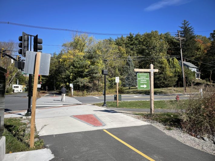 A man crosses a two-lane street at a traffic signal on the course of a paved trail that extends into the woods on the other side. In the foreground a trail sign with a right arrow reads "Town of Sudbury, Sudbury Historic District, Sudbury Town Hall".