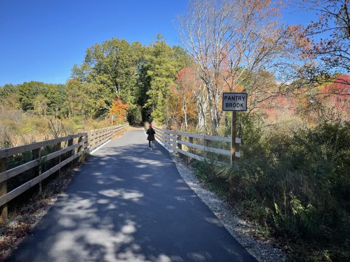 A woman in a black coat and orange hat walks over a short bridge on a straight paved trail lined with wooden fences. The trail runs through a meadow lined with trees with bright red and orange fall leaves. A sign next to the bridge reads "PANTRY BROOK"