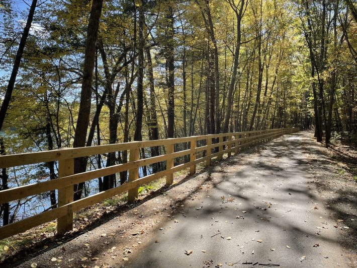 A straight paved trail runs alongside a wooden fence through the autumn woods. To the left, through the trees, a small lake is visible.