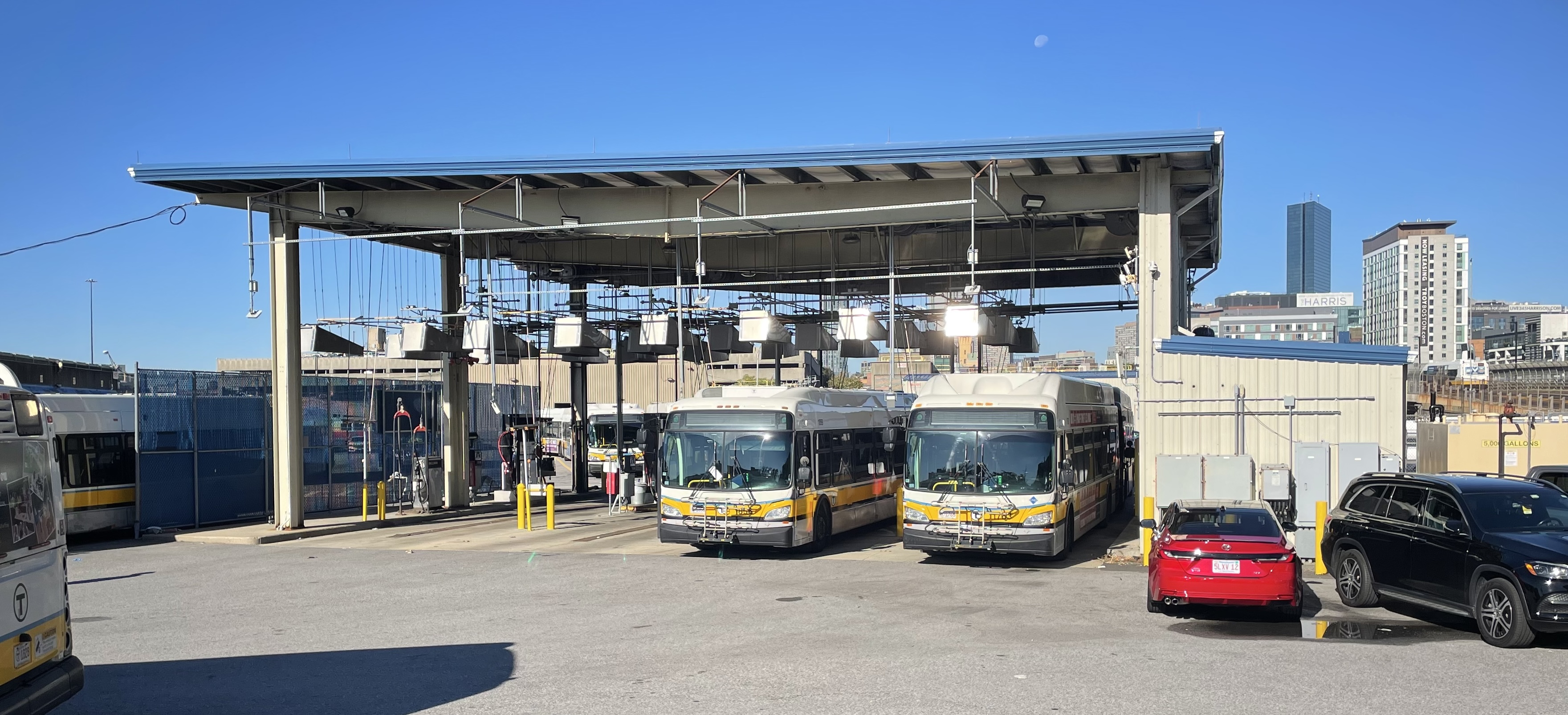Two MBTA buses refueling under a large metal canopy under a blue sky