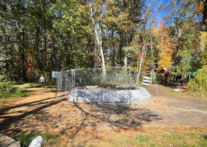 A large circular granite bench sits in the junction of two paved trails surrounded by forests. The granite is surrounded by a chain link fence.