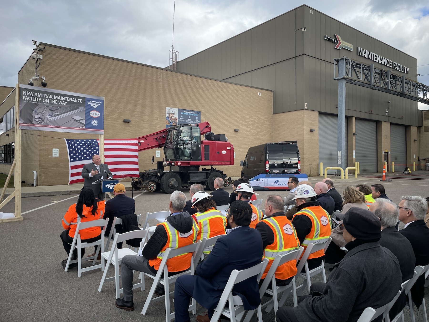 A small crowd seated in folding chairs, wearing overcoats and bright orange construction vests, faces a speaker at a podium in front of a large warehouse building with a sign that says "Amtrak Maintenance Facility." A banner behind the speaker says "New Amtrak Maintenance Facility On the Way". A model of the Acela train is in the background.
