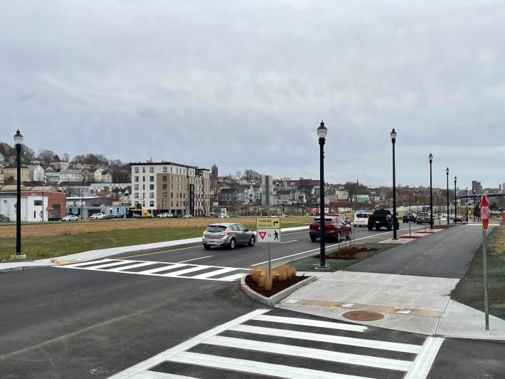 A view down a two-lane, one-way street with a wide shared-use path on the right side and a wide grassy lawn to the left. Across the lawn is another street lined with low-rise buildings, with one taller 6-story building standing out in the middle distance.