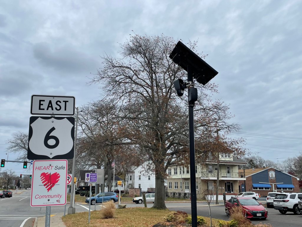A black pole topped with a solar panel and two purse-sized boxes attached just below the panel. To the left is a highway sign that reads "East 6" and below that a sign that reads "HEART Safe Community"