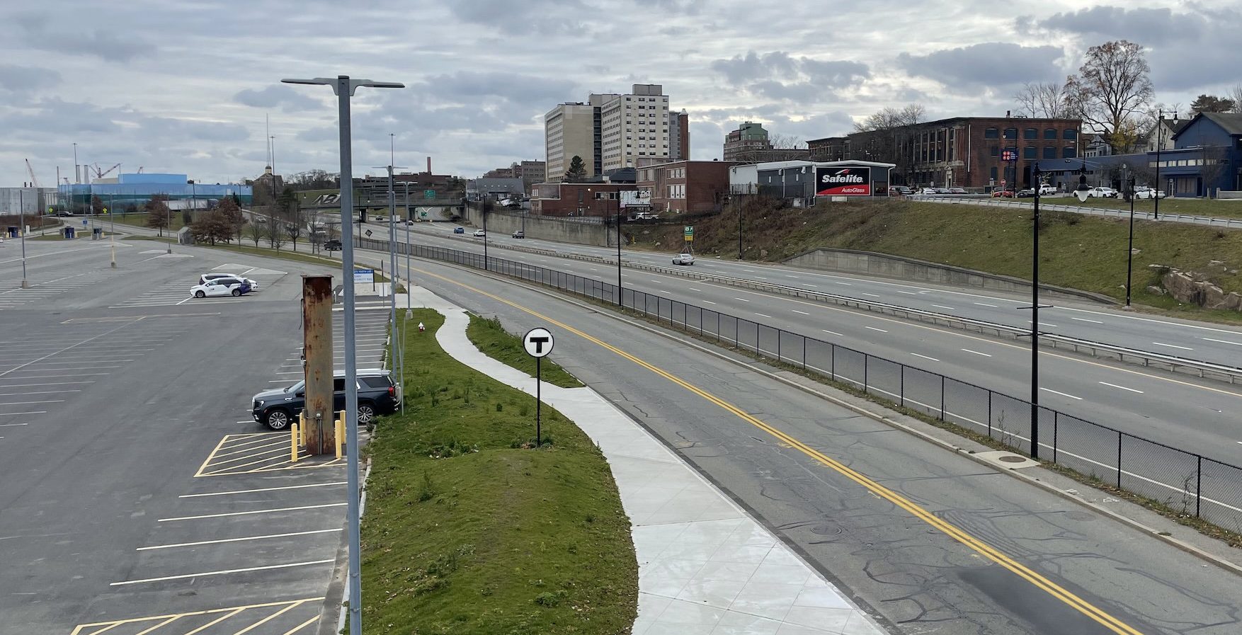 A view from above of a wide 6-lane highway with a two-lane frontage road in the foreground. To the left is a large mostly empty parking lot. In the middle is a newly-built sidewalk, and a sign with the circular MBTA logo. In the distance, on a hillside above the highway, are a handful of taller buildings in a city neighborhood.