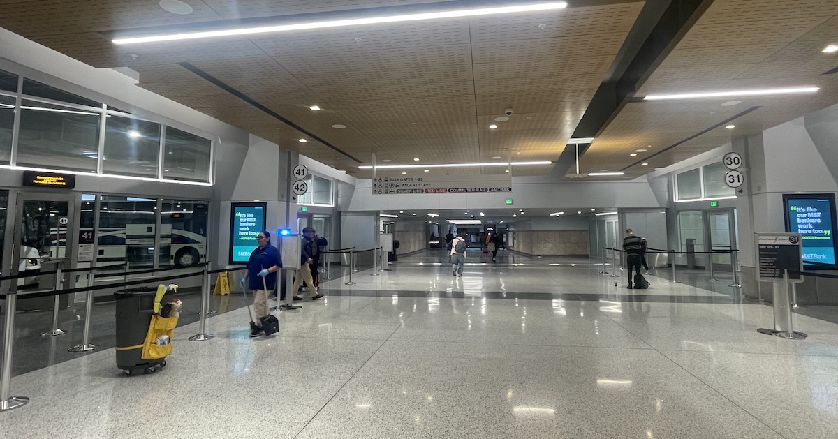 The hall of a bus terminal with polished stone floors and a wood-paneled drop ceiling with long, horizontal lights. In the foreground at left is a cleaning cart, next to several passengers waiting near the door to a platform where buses visible through floor-to-ceiling glass walls are boarding passengers.