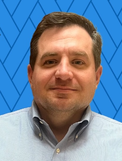 A portrait of a white man with short brown hair and wearing a collared shirt, in front of a blue patterned background. 