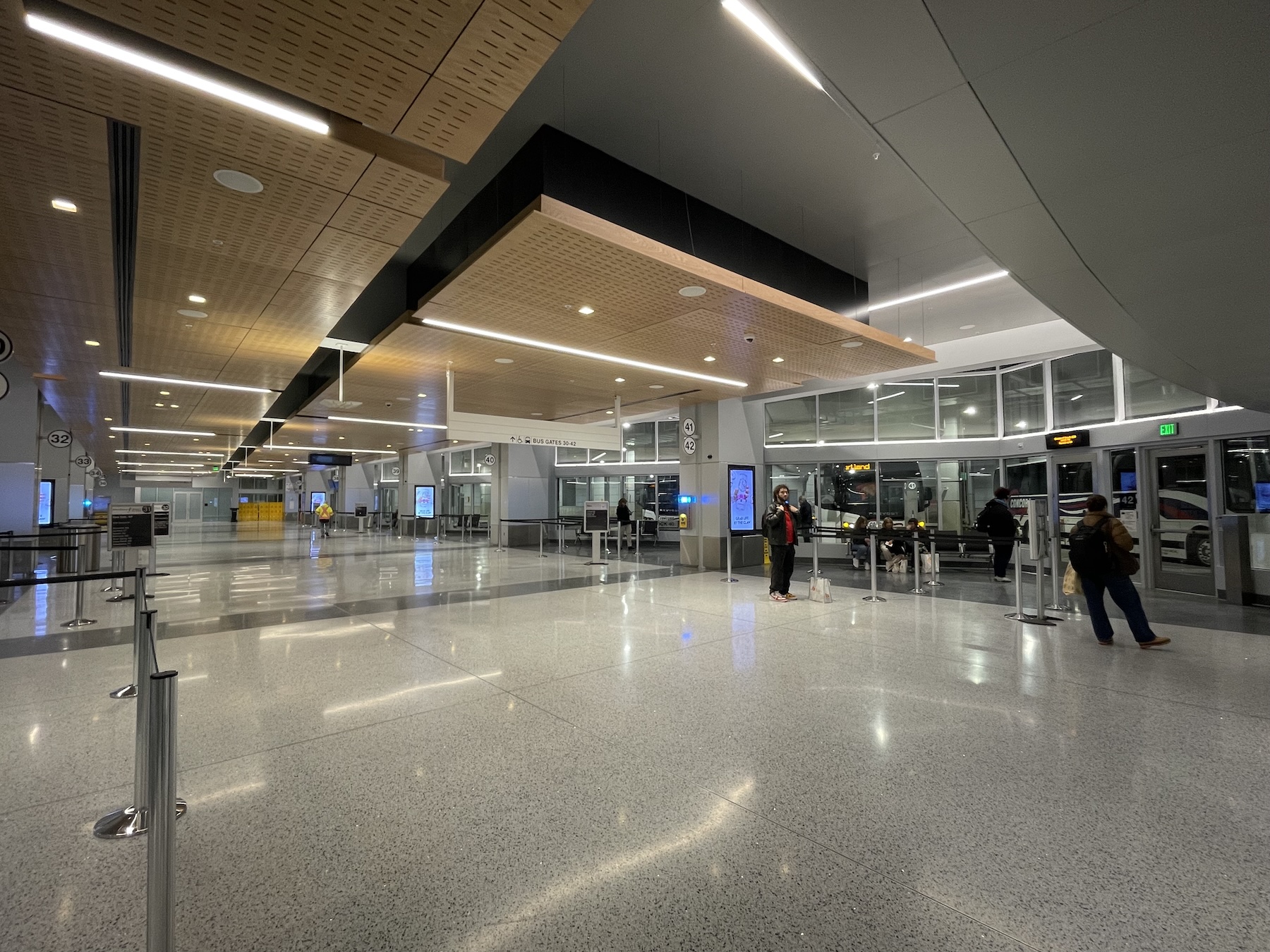 The hall of a bus terminal with polished stone floors and a wood-paneled drop ceiling. In the foreground at right, several passengers wait near the door to a platform where two buses are boarding passengers.