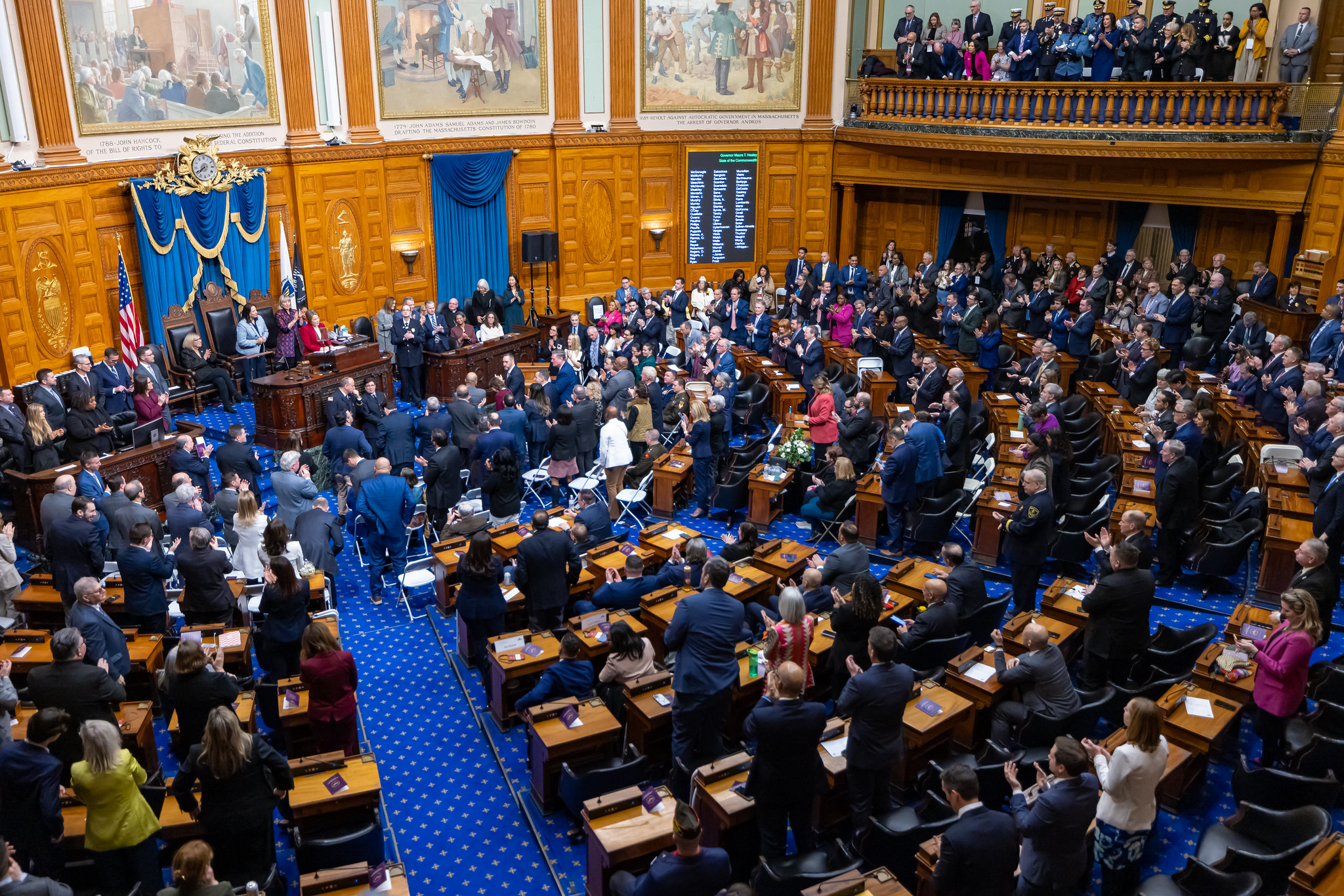 A crowd of people are seated at desks arranged in a semicircle facing a speaker in a red jacket standing at a dias in the front of a large, ornate room.