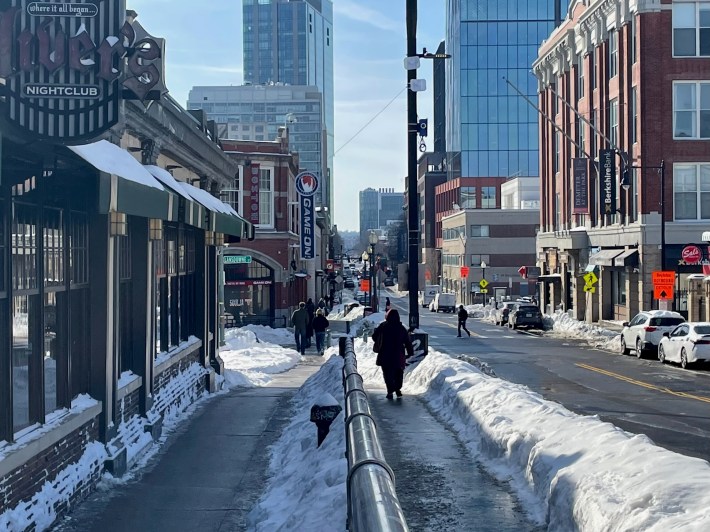 A view down a city street lined with tall buildings in the distance and older mid-rise buildings in the foreground. The street is lined with snowbanks and the sidewalks and street itself are cleared of snow.