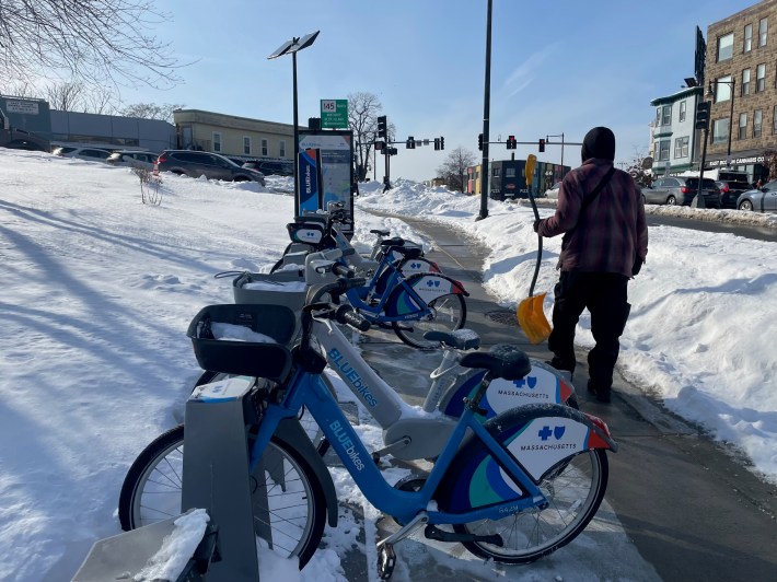 A man with a shovel walks past a Bluebikes bikesharing station, which sits on a sidewalk in between two tall snowbanks. 