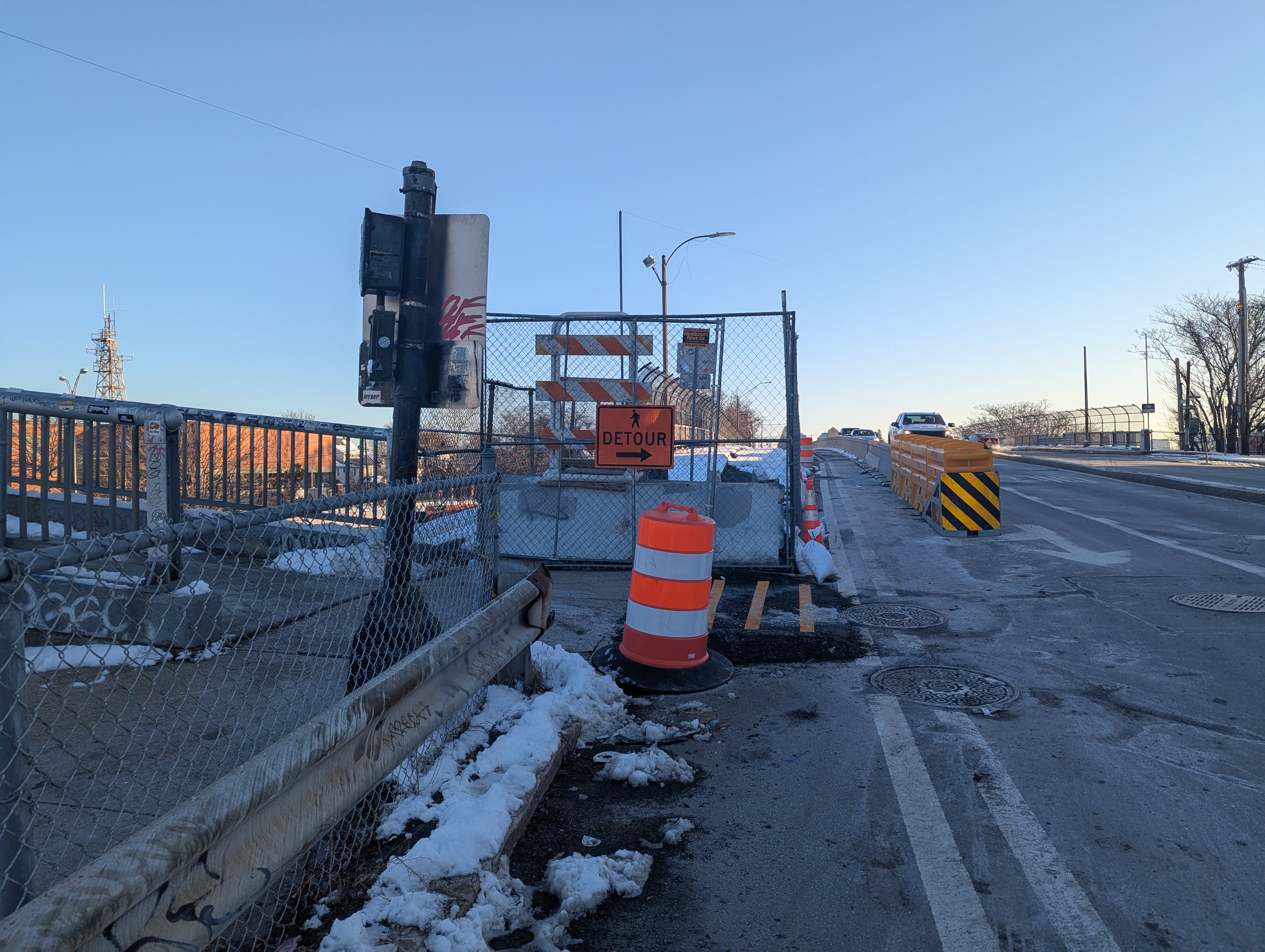 A photograph of a bridge construction zone in winter. In the foreground a metal crash barrier and chain link fence separate a concrete sidewalk (left) from a multi-lane roadway (right). In the middle distance another fence blocks the sidewalk, with an orange "detour" sign pointing right towards the roadway behind an orange construction barrel. In the distance, a crash barrier in the roadway separates car traffic from a temporary sidewalk across the bridge. On the left edge of the photo a second pedestrian bridge joins with the sidewalk.