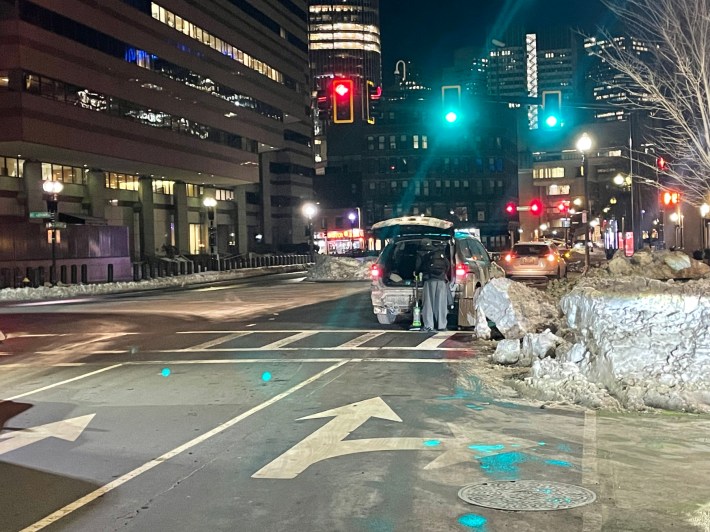 A wide city street at night, with a snow pile blocking the right-most driving lane and bike lane. A car with its trunk open is stopped in the roadway under a green traffic light next to the snow pile. 