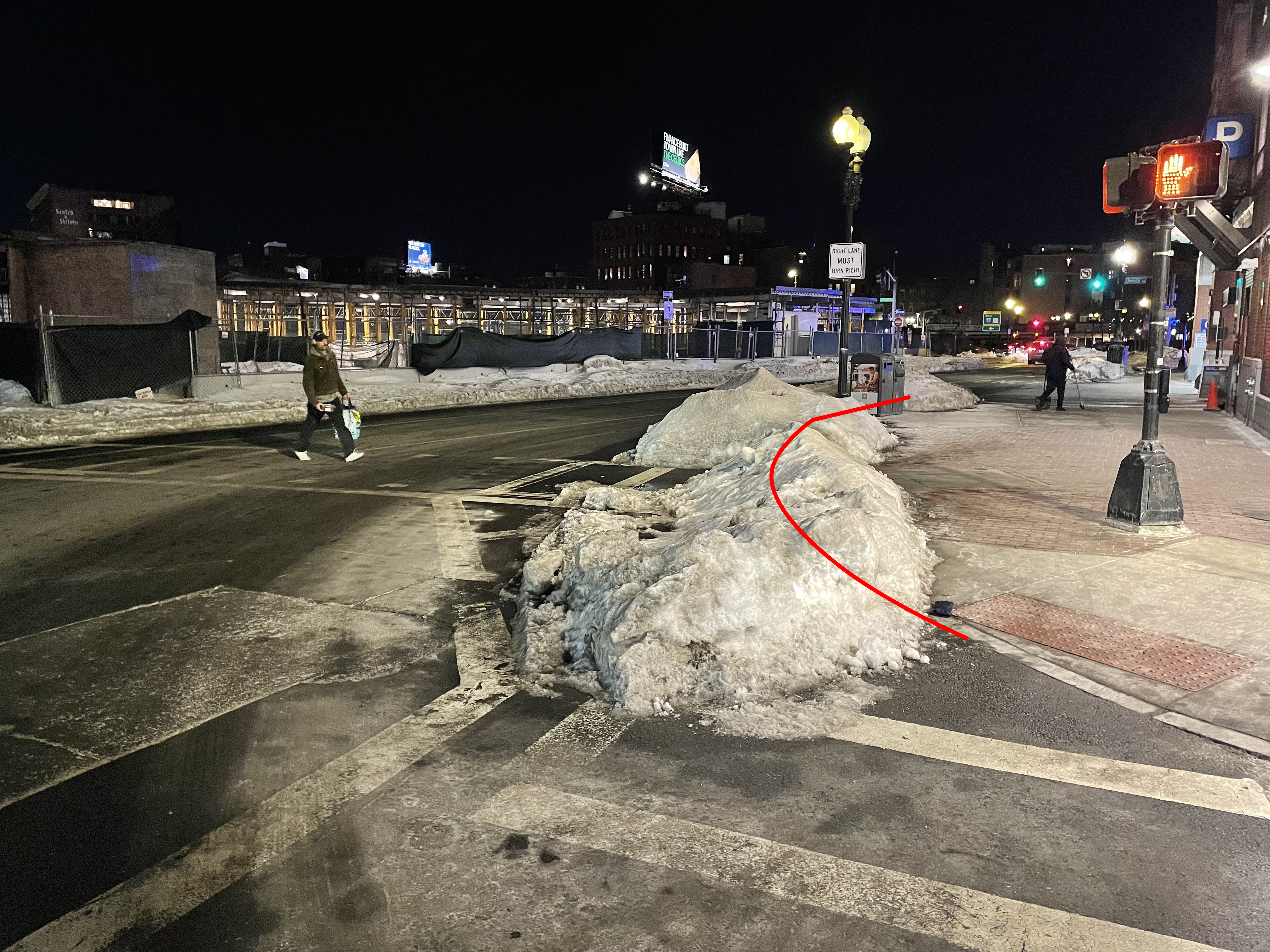 A large snowbank blocks the edge of a roadway on a street corner at night. A red line through the middle of the snowbank indicates the approximate location of a buried curb line. To the left, a pedestrian in a crosswalk approaches the snowbank.