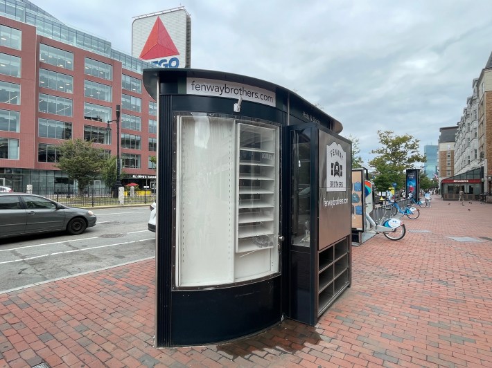 An empty retail kiosk sits on a brick sidewalk in Kenmore Square. Above the newsstand, the neon Citgo sign is visible on the other side of the street.