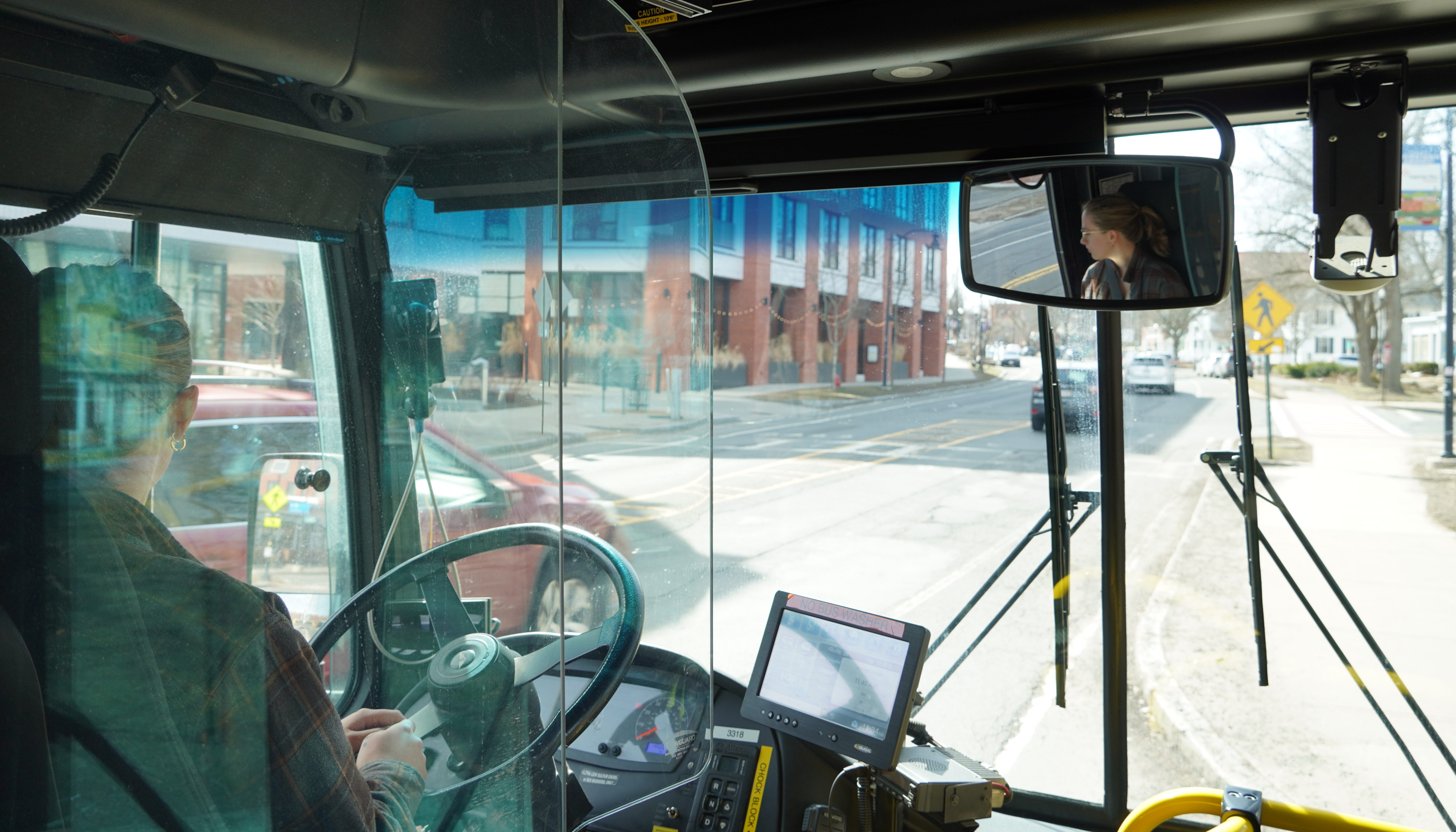 A photo of UMass Transit employee Ash Larner driving a city bus don a downtown street. The photo is taken from behind the driver's seat of a city bus, with Larner's face visible in profile the rear-view mirror at upper right.