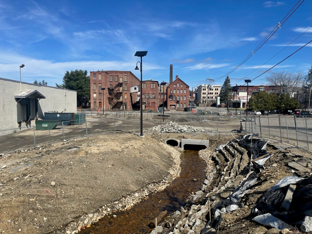 A stream flows through a rocky channel through the middle of a construction site. Chain-link fencing surrounds the steep dirt banks that descend about 10 feet down from the level of the parking lot that surrounds the stream. In the distance is a cluster of old brick multi-story mill buildings, mostly boarded up, and a cinder-block warehouse building to the left.