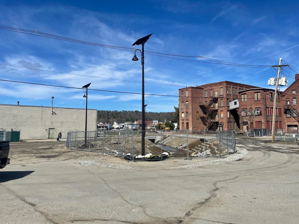 A construction site in the middle of a large, old asphalt parking lot. In the center of the photo, chain-link fencing surrounds the steep dirt banks that descend about 10 feet down from the a stream that flows into a culvert pipe in the distance. To the right is a cluster of old brick multi-story mill buildings, mostly boarded up, and there's a cinder-block warehouse building to the left.