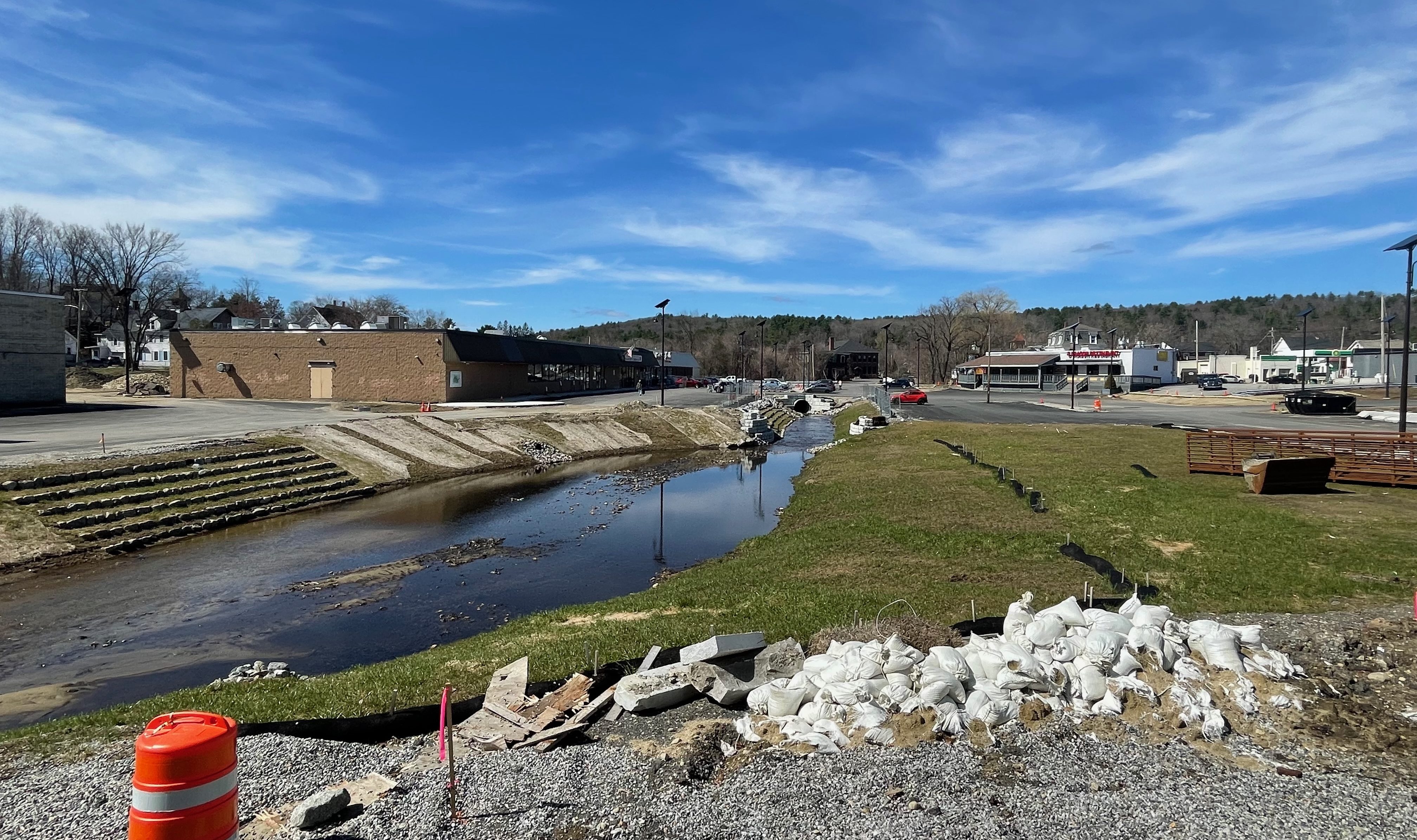 A construction site around a broad, shallow stream. The banks of the stream are a mix of terraced rip-rap stones to the left and a gently-sloping lawn to the right with sandbags and an orange construction barrel in the foreground. In the distance is a single-story strip mall to the left and a surface parking lot to the right.