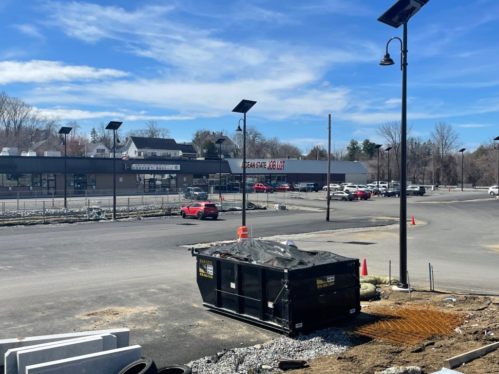 A freshly-paved parking lot in front of an old strip mall with a sign that says "OCEAN STATE JOB LOT". In the middle of the parking lot chain link fencing surrounds a construction area, and in the foreground is a large construction dumpster surrounded by crushed stones and new granite curbs.