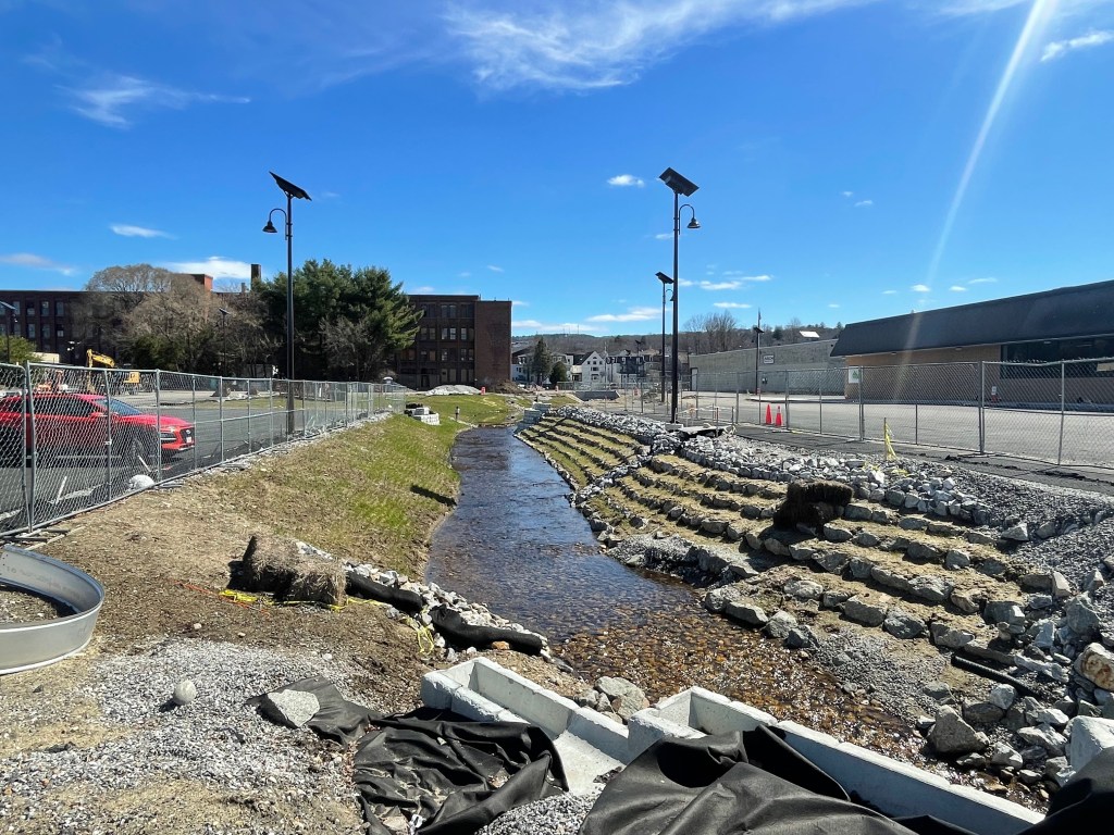 A construction site around a broad, shallow stream. The banks of the stream are a mix of terraced rip-rap stones to the right and a sloping lawn to the left. On the tops of each bank is a chain link construction fence and several tall lampposts topped with solar panels. Beyond the fencing is a freshly-paved parking lot.