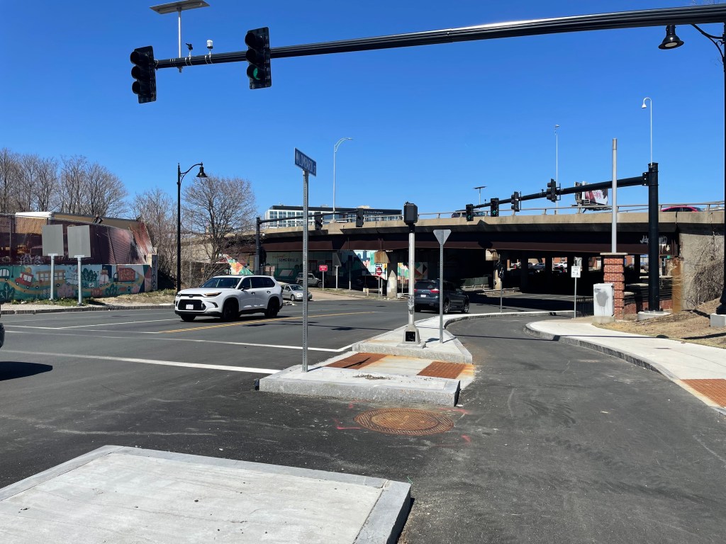 An intersection with several traffic lights and curbs. A median in the center of the photo divides a bike path (center right) from a three-lane roadway (left). In the distance a highway overpass flies over the bike path and the street.