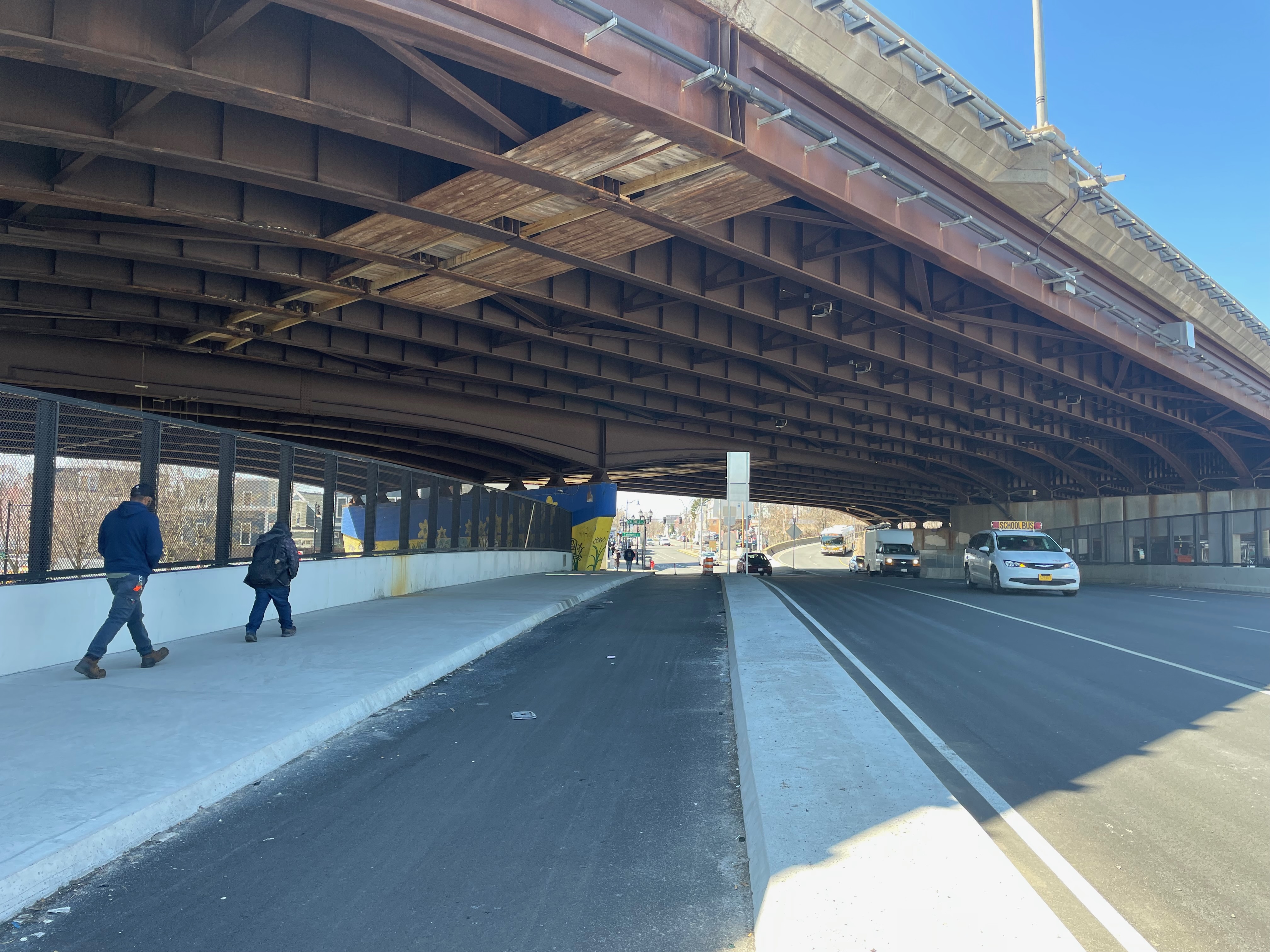 A wide roadway shaded under a huge brown highway overpass under a blue sky. At left, several pedestrians walk along a new concrete sidewalk on the side of the road. In the center is a concrete curb that separates a bike lane from the motor vehicle lanes at right.