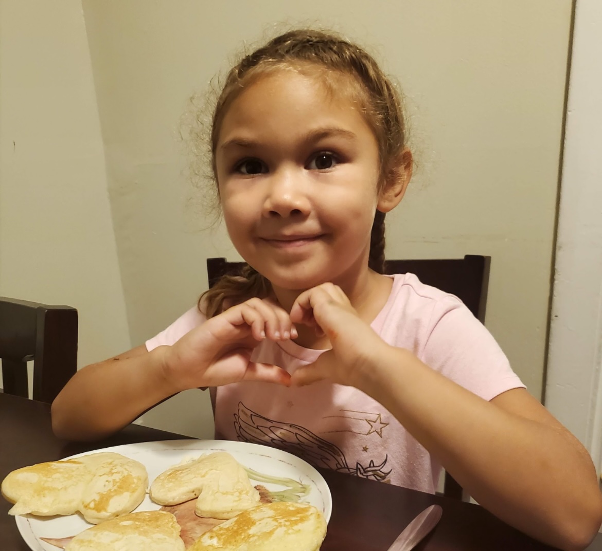 A grinning brown-skinned girl makes a heart shape with her hands while sitting in front of a plate of pancakes.