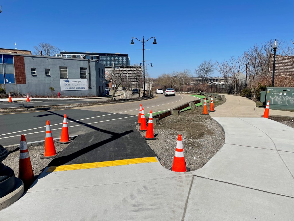 Several orange construction cones line a new asphalt ramp from the sidewalk to a bike lane in the adjacent roadway. On the right edge of the photo, a new concrete sidewalk connects to an older sidewalk segment in the middle distance. A high-rise office building is visible on the horizon.