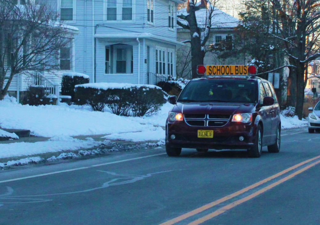 A dark red school transportation van with a yellow "SCHOOL BUS" sign on top of the van with red lights on either side drives down a street with snow and houses.