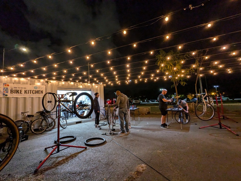 A nighttime scene showing people repairing bicycles outside a white shipping container with rows of warm, glowing string lights above.