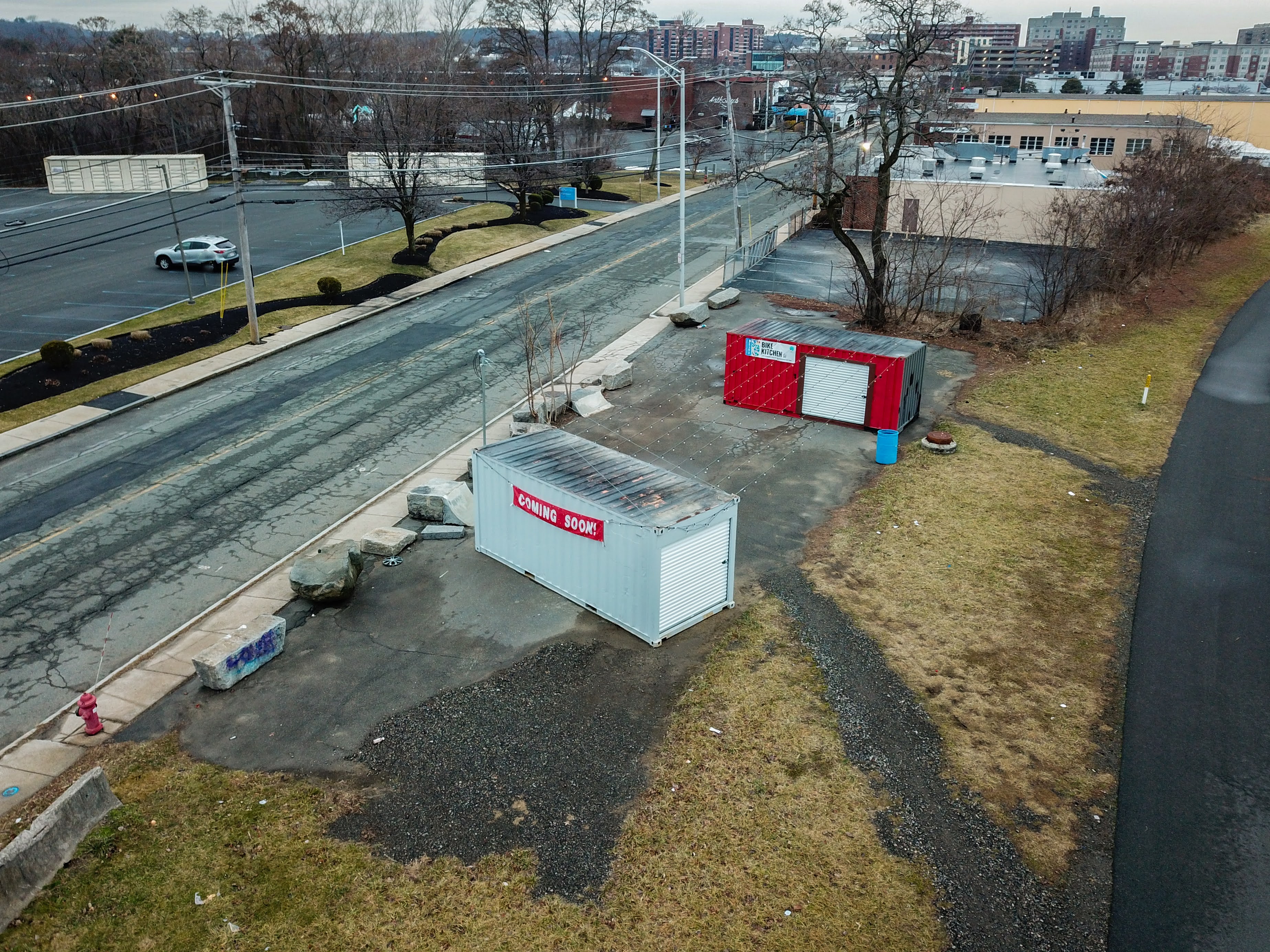 An aerial view of a roadside lot containing two shipping containers, one red and one grey, with a red "Coming Soon!" banner.